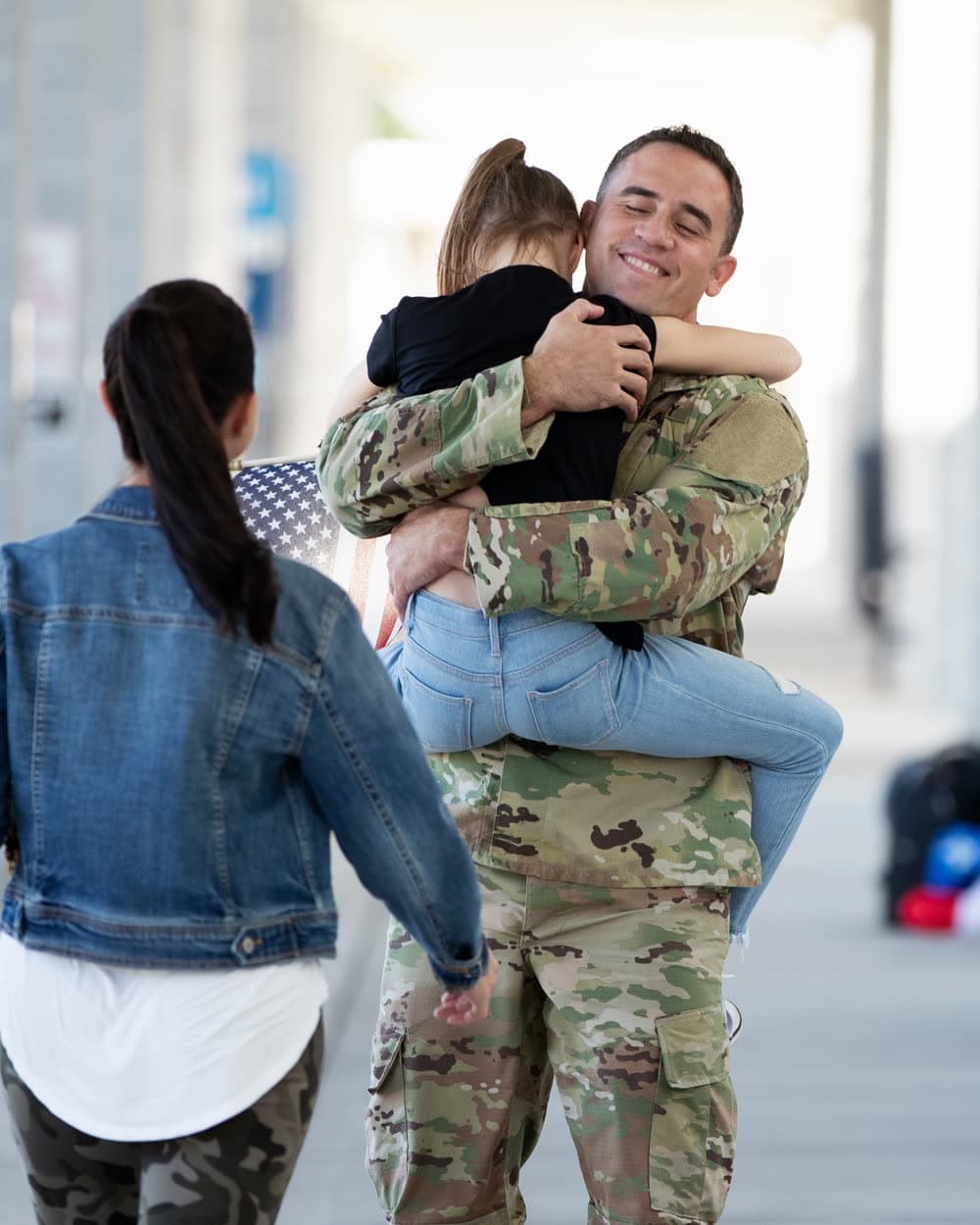 A service member in uniform holding their child at a homecoming, family walking to meet them.