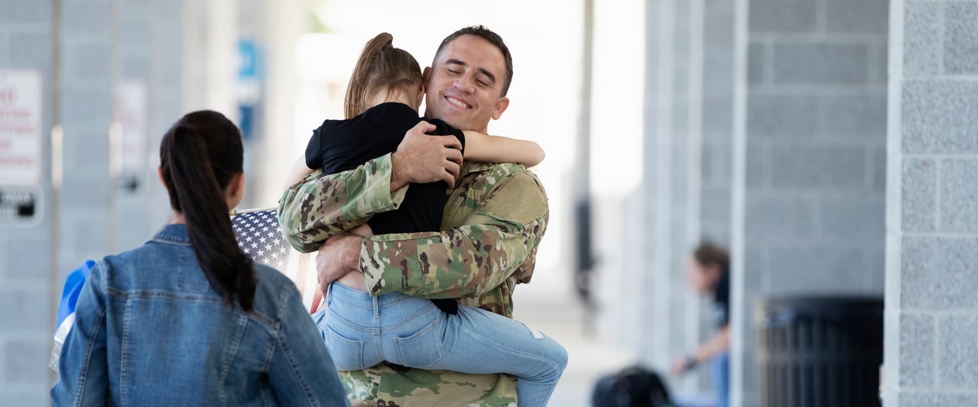 A service member in uniform holding their child at a homecoming, family walking to meet them.